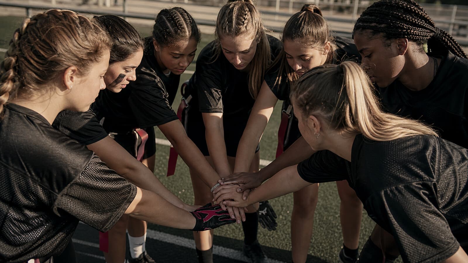 Seven teenage girls in a tight sideline huddle, hands stacked center.