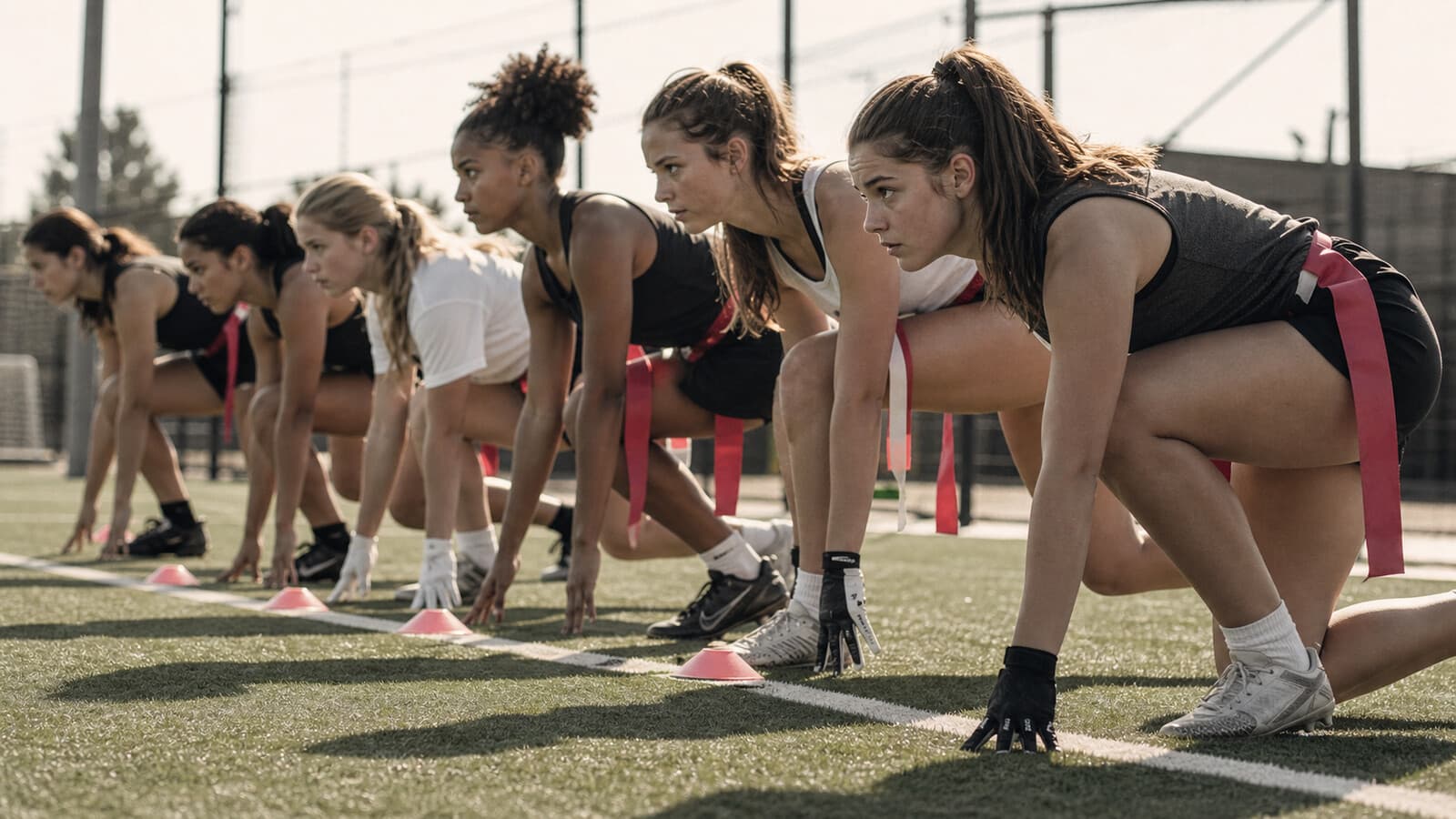 A line of teenage girls in flag football gear in a three-point sprint stance, captured from a low angle.