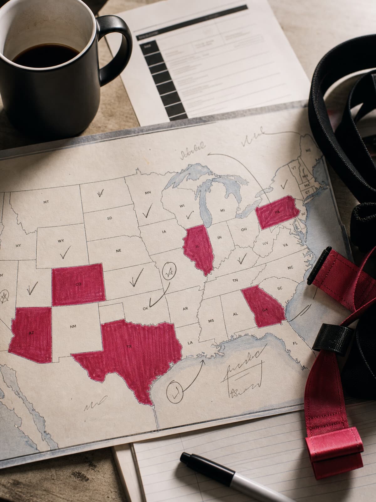 An overhead view of a printed US map with several states marked in dusty rose, a coffee mug, a flag football flag belt, and handwritten notes.