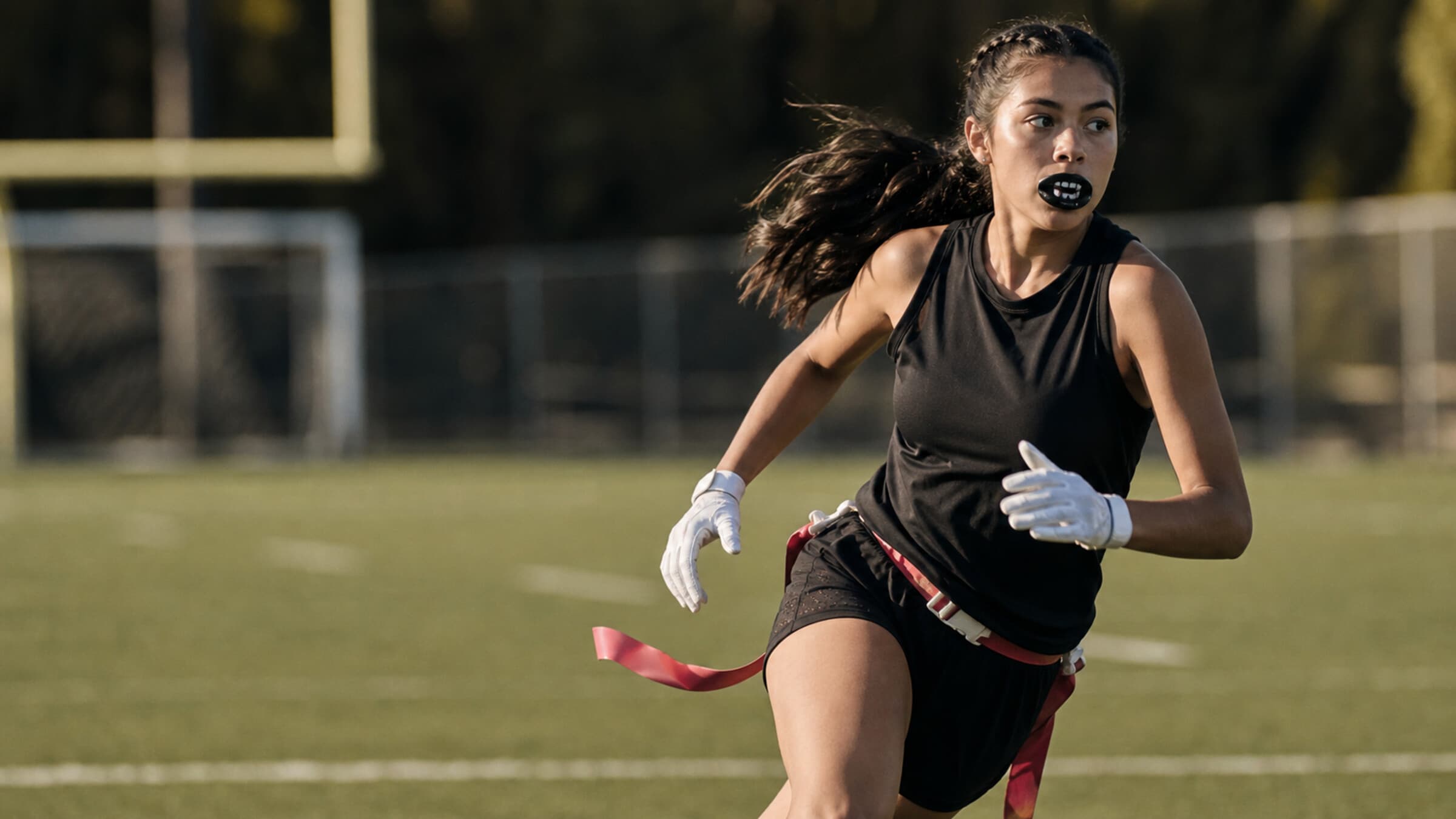A girls flag football QB in mid-stride on a turf field, flag belt visible, mouthguard out, focused downfield.
